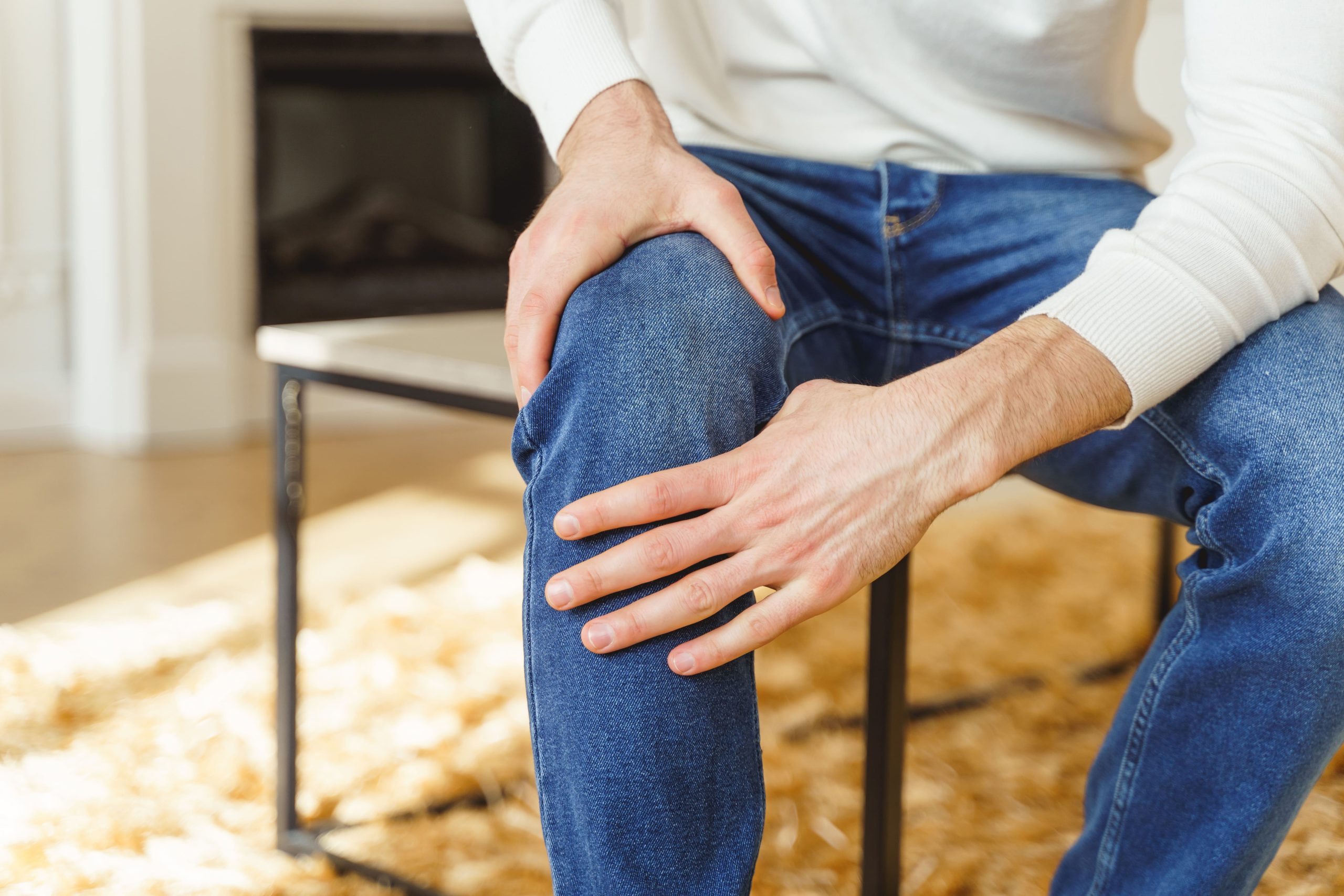 An adult seated in a home interior, holding their knee in discomfort.