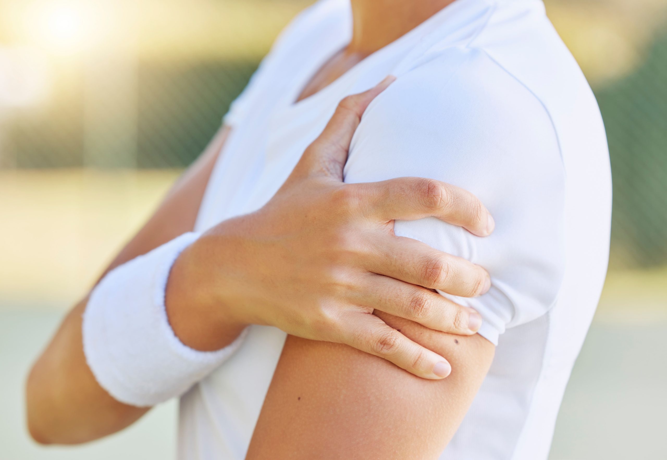 An adult holding their shoulder in discomfort while wearing a white shirt and a wristband, likely outdoors.