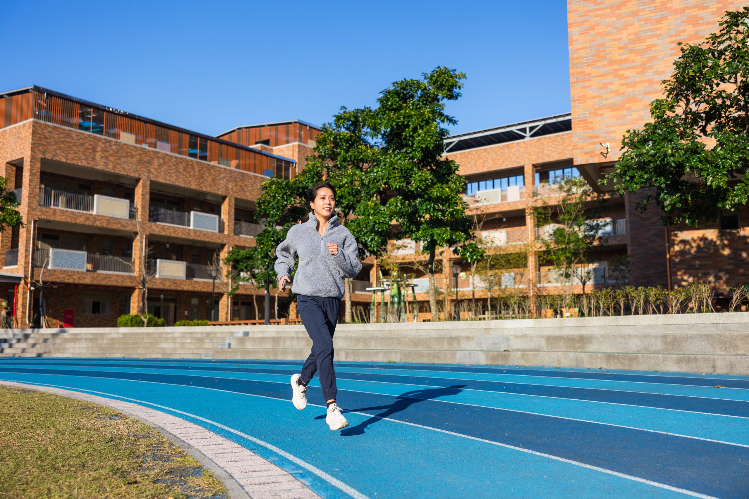 A person jogging on a blue running track under a clear blue sky with modern buildings in the background.