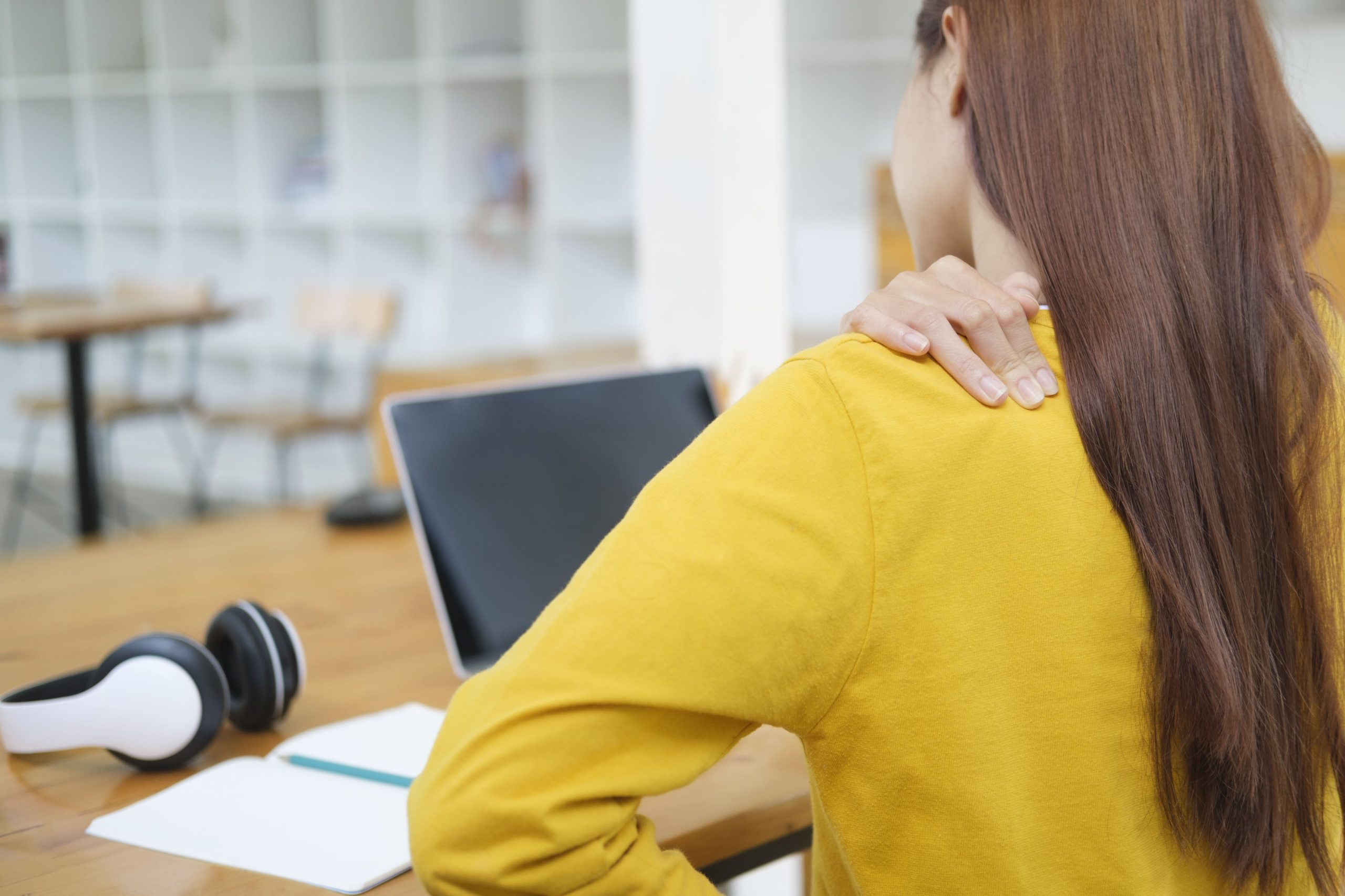 A young woman in a yellow shirt feeling back pain while sitting at an office desk with a laptop and headphones.
